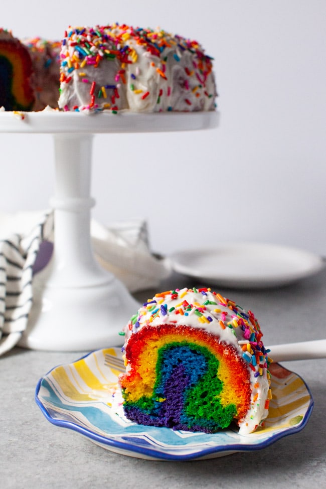 Slice of rainbow cake on a plate next to tray of cake