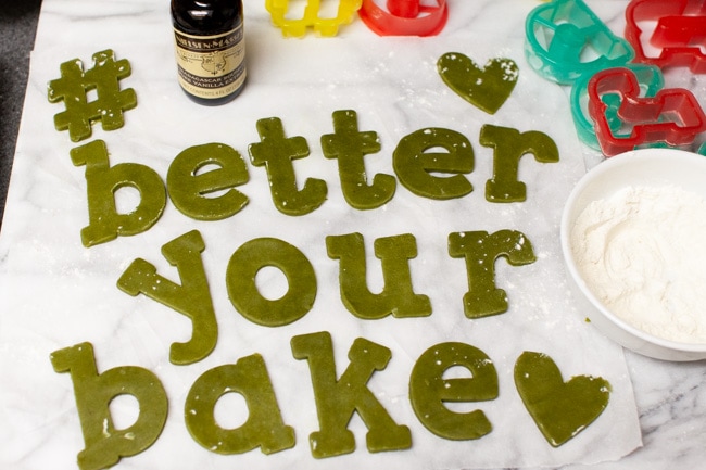 Matcha Cookie dough cut to spell #betteryourbake with bottle of vanilla, cookie cutters, and bowl of flour in background from thelittlekiitchen.com