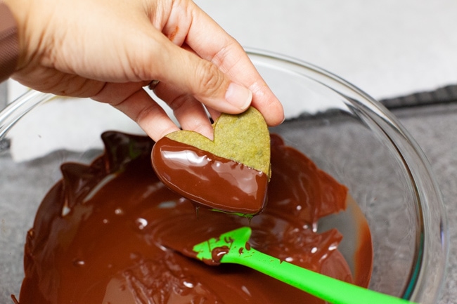 Hand dipping heart-shaped Matcha Cookies into glass bowl of melted chocolate from thelittlekiitchen.com