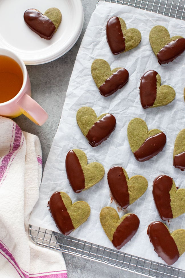 Chocolate Dipped Heart Shaped Matcha Cookies on parchment paper and cooling rack with kitchen towel, pink cup of tea, and cookie on plate in the background from thelittlekiitchen.com