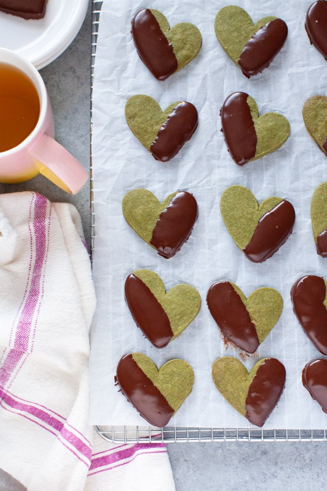 Heart Shaped Chocolate Dipped Matcha Cookies on white parchment paper, next to kitchen towel, cup of tea, and white plate from thelittlekiitchen.com
