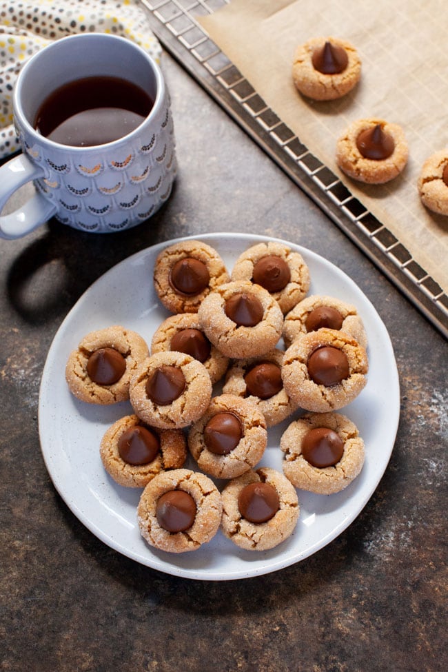 Peanut Butter Blossom Cookies from thelittlekiitchen.com Peanut Butter Cookies topped with a Hershey Kiss sit on a off-white plate next to a cup of coffee.